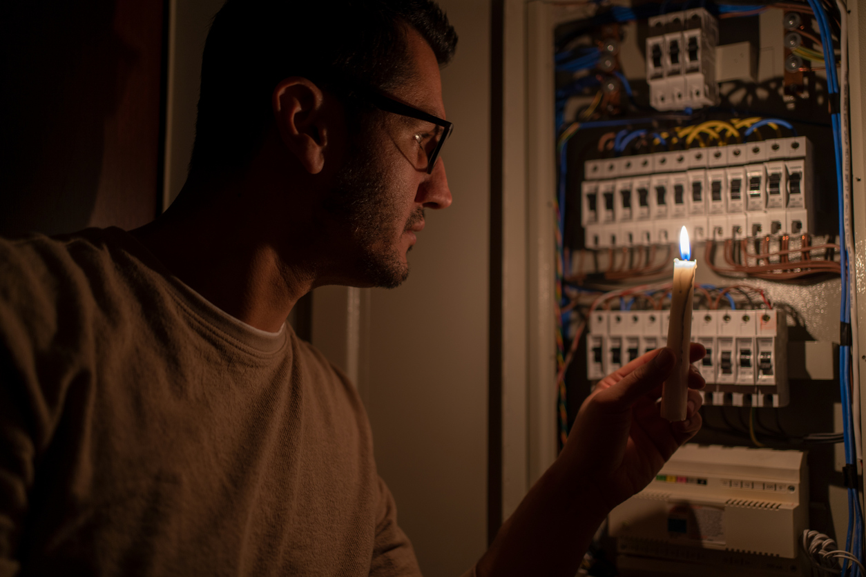 Man Checking Home Fuse Box By Candlelight During Power Outage